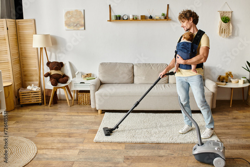 bearded man with infant baby boy in carrier vacuuming apartment at home, fatherly love and housework photo