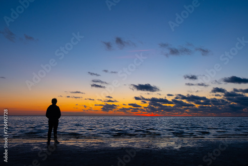 This tranquil image captures the silhouette of a lone individual standing on the shore, immersed in the serenity of the twilight. The horizon is painted with warm hues of orange and yellow, fading