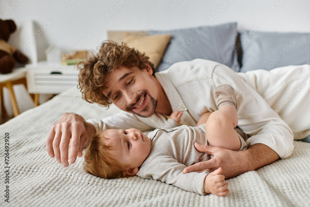 cheerful father with curly hair and beard looking at his infant baby son on bed, precious moments cheerful father with curly hair and beard looking at his infant baby son on bed, precious moments