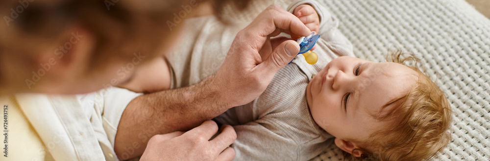 Fototapeta premium top view of curly father giving pacifier to his infant baby son on the bed, fatherhood banner