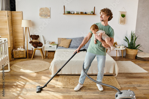 happy man multitasking housework and childcare, father vacuuming bedroom with infant boy in arms photo