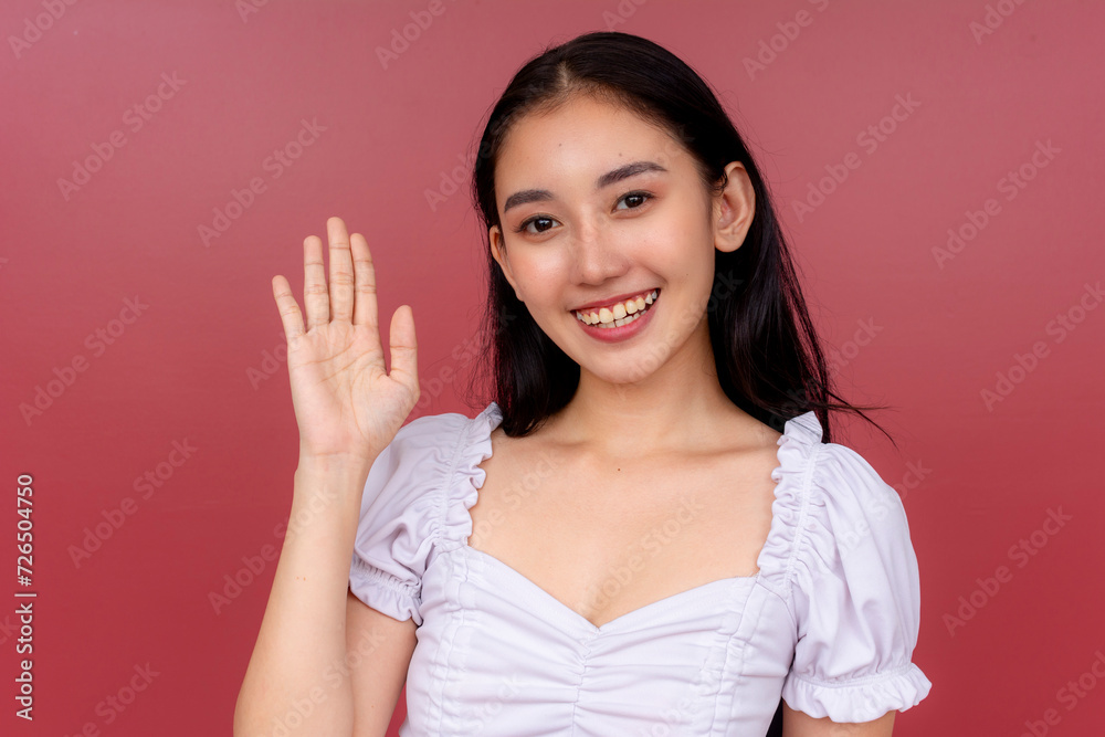 A happy and congenial Asian woman in a white blouse greets by waving her hand against a vibrant red background, exuding friendliness. A happy and congenial Asian woman in a white blouse greets by waving her hand against a vibrant red background, exuding friendliness.