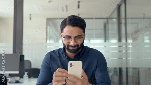 Busy Indian business man worker using cell phone at work in office. Professional businessman entrepreneur working on cellphone, smiling male employee executive holding smartphone checking mobile apps. photo