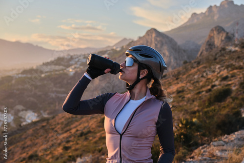 Woman cyclist in cycling kit and a helmet is drinking water from a sports bottle at sunset on mountains background. Cyclist silhouette. 