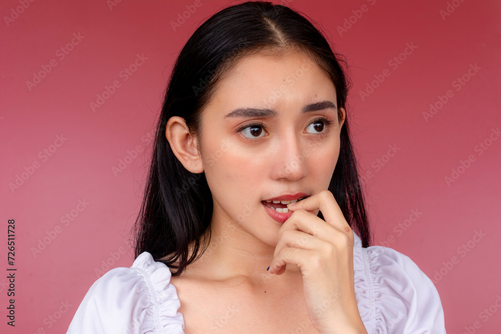 Portrait of an anxious young Asian woman with her hand on her lips, worrying about her situation. Isolated over a vibrant red background. Portrait of an anxious young Asian woman with her hand on her lips, worrying about her situation. Isolated over a vibrant red background.
