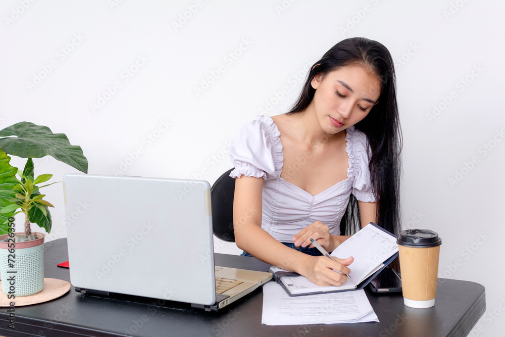 A dedicated and studious young woman is engaged in learning, writing notes next to her laptop and coffee at a study desk. A dedicated and studious young woman is engaged in learning, writing notes next to her laptop and coffee at a study desk.