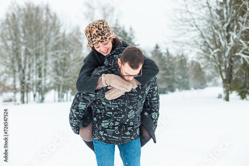 beautiful couple of lovers are having fun together in a winter snowy park. A handsome man carries on his back a beautiful girl with a smile outdoors