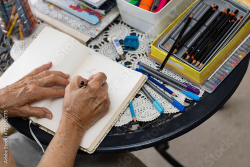 elderly wrinkled hands holding a pencil and writing in a notebook with drawing supplies on table photo
