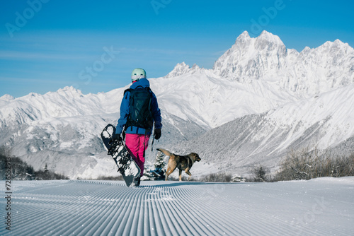 Female snowboarder walking with dog on Snow trail from ratrak preparation, freshly groomed ski slope in high mountains ski resort