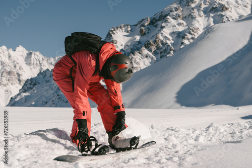 woman snowboarder wearing red ski suit and helmet fastens snowboard bindings while standing on top of slope in ski resort. Against background of grandiosely beautiful mountain range