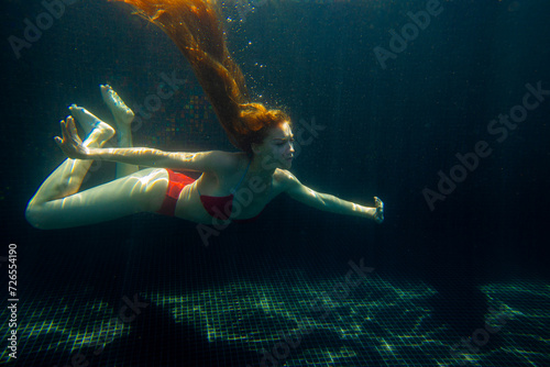 A beautiful sporty girl poses underwater with loose hair against the bright rays of the sun from the surface.