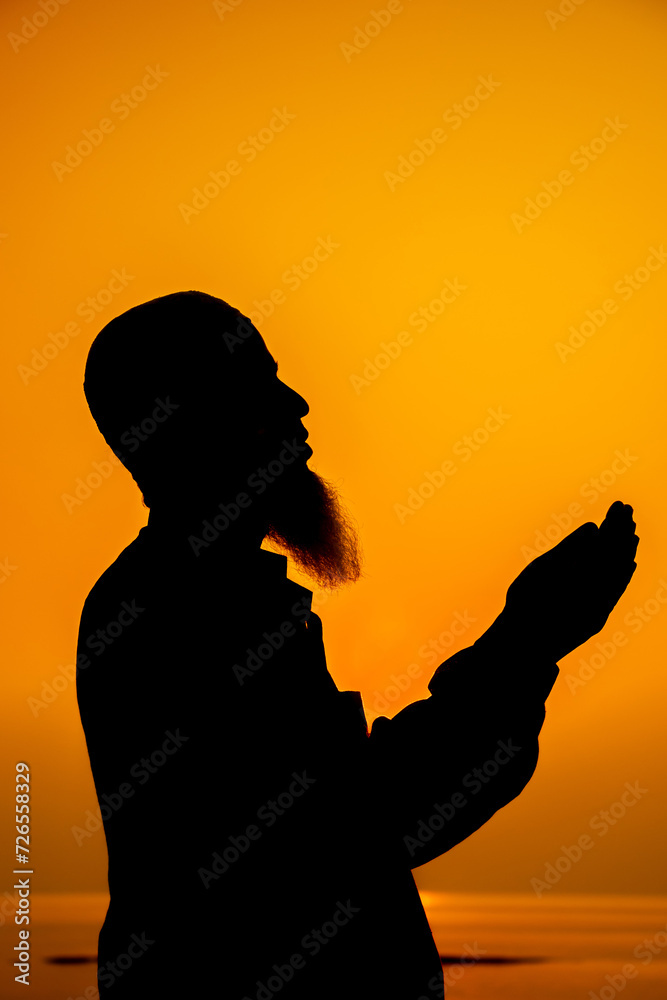 Asian Muslim bearded man praying with hands raised to God on sunset background. Silhouette of an old age Muslim praying during sunset.