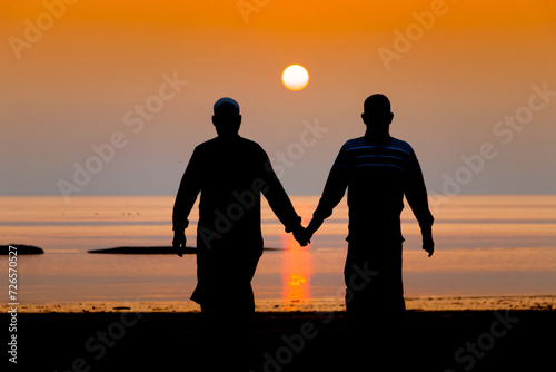 Silhouette view of two Muslim men walking hand in hand on a sea beach during twilight sunset. Friendship is an immortal concept even in old age.