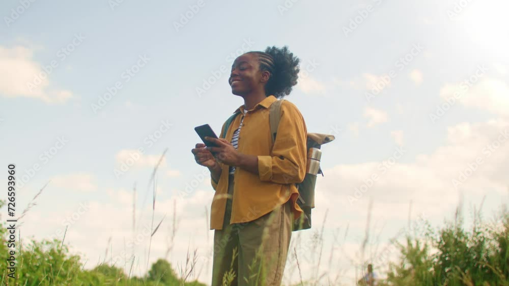 Pretty African American woman with backpack standing on mountain or field. Chatting with someone on her smartphone. Having good mood while messaging on social media. Holiday concept. Pretty African American woman with backpack standing on mountain or field. Chatting with someone on her smartphone. Having good mood while messaging on social media. Holiday concept.
