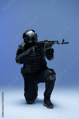 Soldier in black military uniform for city with helmet, action camera and machine gun, photo in studio. Army man stands on one knee, defensive position