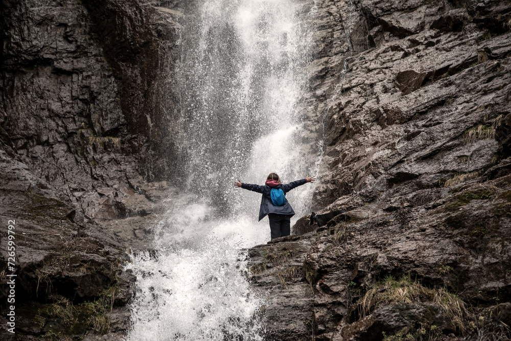 Obraz premium A girl and a dog stand against the backdrop of a waterfall. Beautiful nature. Splashes of water. American Staffordshire Terrier. Mountains.