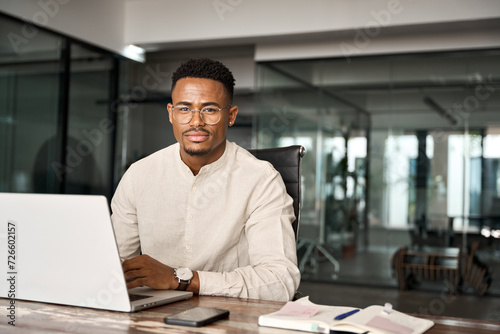 Confident young professional African American business man looking at camera sitting at work desk using computer. Portrait of male company employee or entrepreneur with laptop in office, copy space.