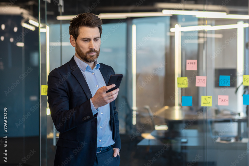 Successful and confident male businessman standing inside office in suit, holding hand in pocket and using mobile phone, reading and sending message, calling, waiting for meeting. Successful and confident male businessman standing inside office in suit, holding hand in pocket and using mobile phone, reading and sending message, calling, waiting for meeting.