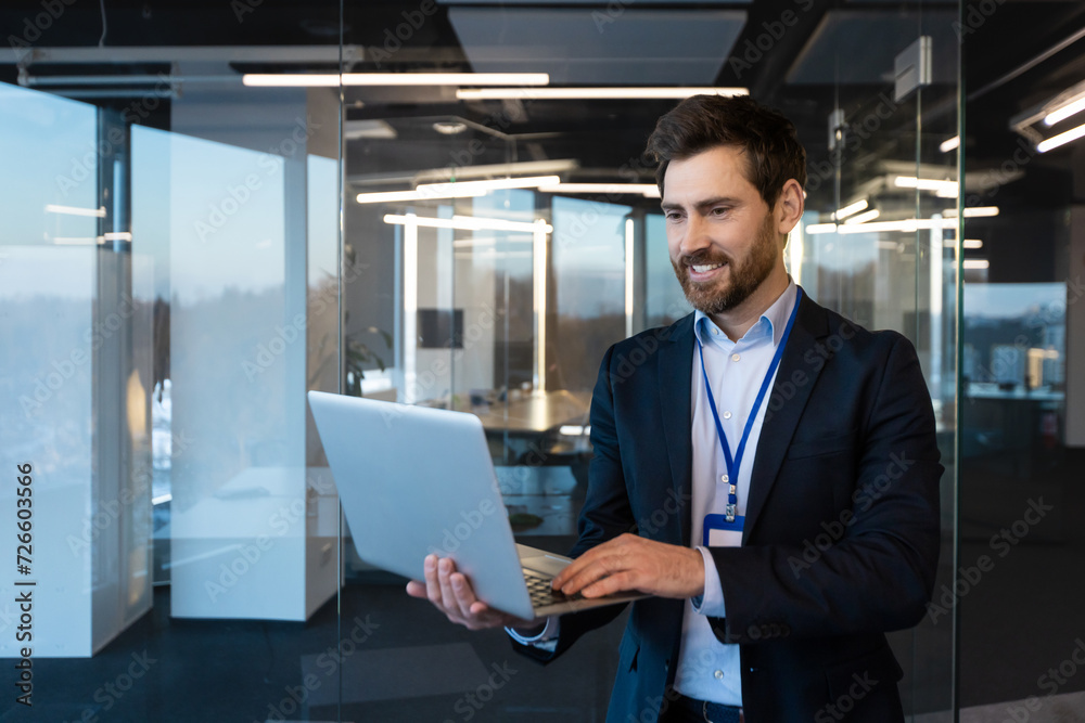 Fototapeta premium Close-up photo of a smiling young businessman. a specialist programmer standing in a suit and with a badge in the office at a conference, holding and using a laptop.