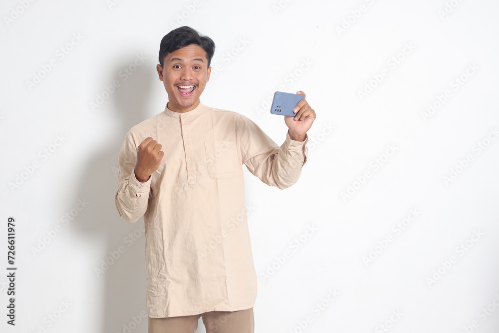 Portrait of excited Asian muslim man in koko shirt holding mobile phone and playing games on his smartphone, raising hands, celebrating success, become a winner. Isolated image on white background Portrait of excited Asian muslim man in koko shirt holding mobile phone and playing games on his smartphone, raising hands, celebrating success, become a winner. Isolated image on white background