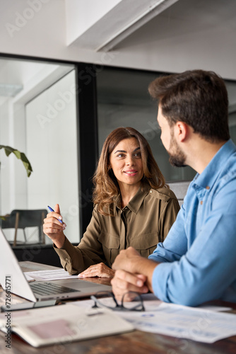 Two busy professional business people working in office with laptop computer. Middle aged female executive manager talking to male colleague having conversation sitting at workplace. Vertical