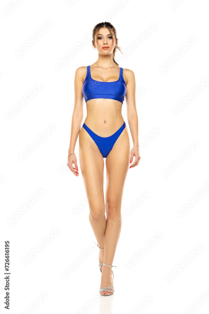 Young beautiful brunette woman in blue bikini swimsuit walking on a white background. Young beautiful brunette woman in blue bikini swimsuit walking on a white background.