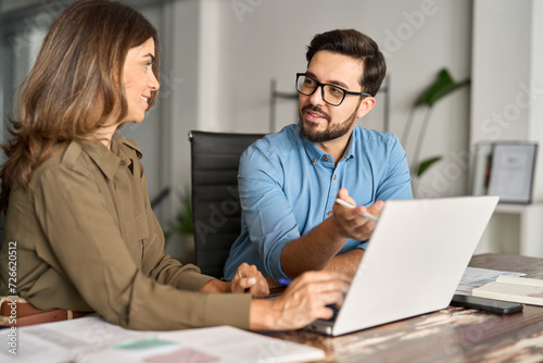 Two busy professional business people working in office with laptop computer. Mid aged mature Latin female executive manager talking to male colleague having conversation sitting at workplace.