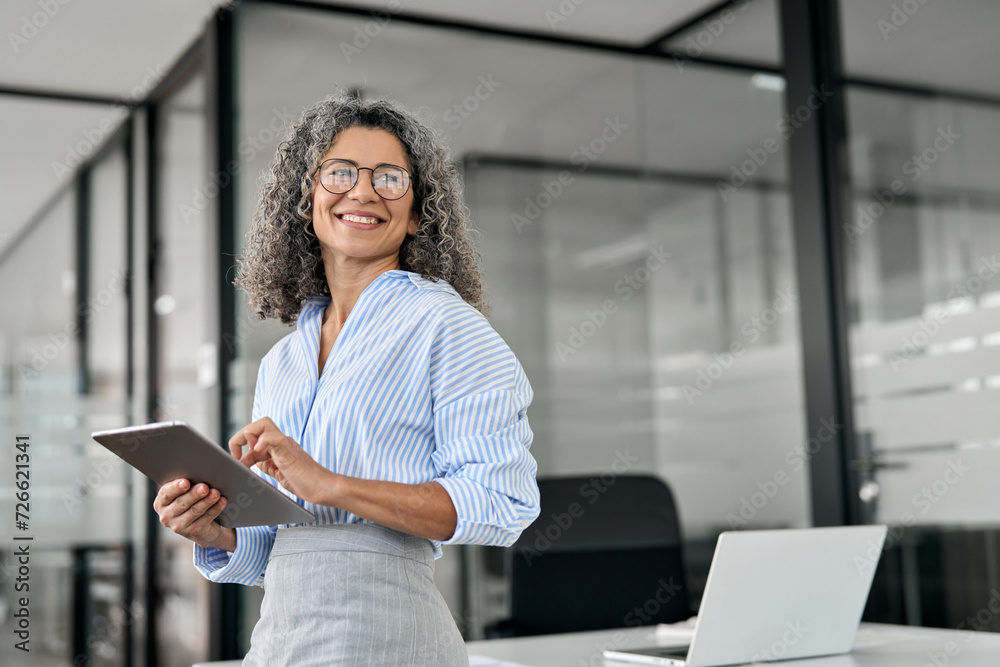 Smiling mature professional business woman bank manager, older happy female executive or lady entrepreneur holding digital tablet pad standing in office at work, looking away at copy space. - obrazy, fototapety, plakaty 
