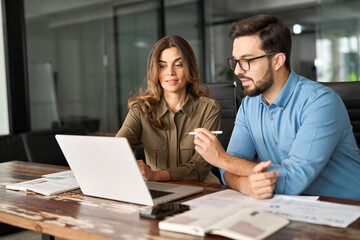 Two busy colleagues working together talking using laptop in office. Middle aged Latin female manager teaching young male worker looking at computer discussing business plan at office meeting.
