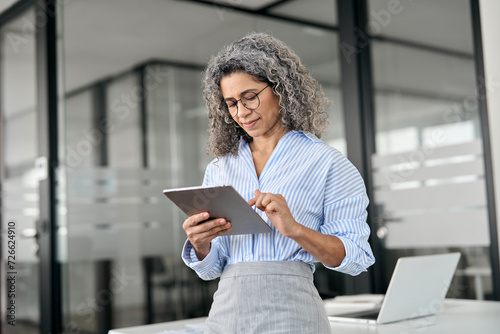 Busy middle aged professional business woman using tab computer in office. Mature senior businesswoman bank manager, older female corporate executive holding digital tablet standing at work.