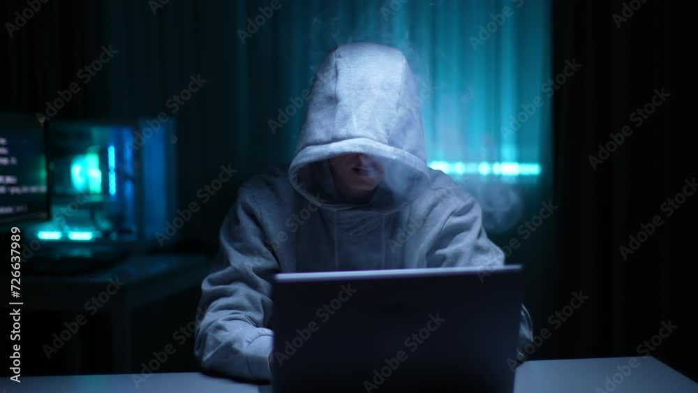 Unrecognizable hacker man in hoodie vaping electronic cigarettes and hacking online web site or engaging hacking into security systems at dark room. Hooded freelancer working laptop computer from home