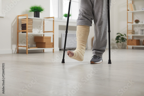 Close up photo of a handicapped man with broken leg trying to walk using his crutches at home. Disabled guy with plastered leg in rehabilitation and recovery having fractured limb. photo