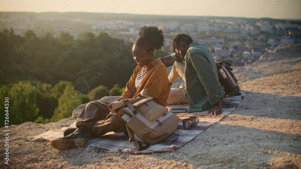 Warm view of African American couple sitting on edge of mountain and discussing something. Looking for something with help of their binoculars. Pointing their fingers at some location.