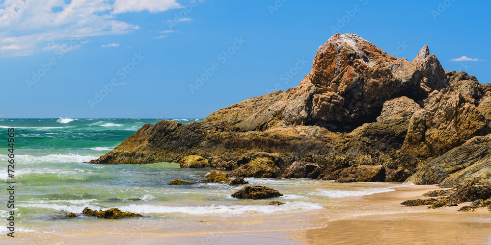 Australian coast with volcanic rocks on the shore, view from the beach to the horizon with blue water and waves breaking on the rocks, summer sunny day.