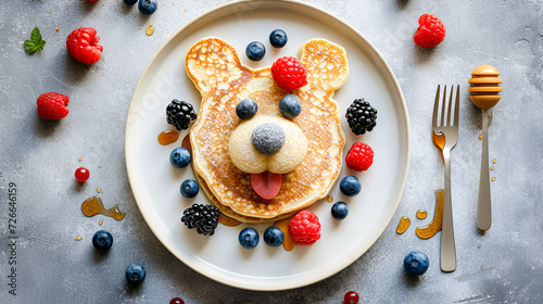 children's breakfast pancakes in the shape of a cute bear's face with berries and honey on a light gray concrete background, top view with copy space for recipe