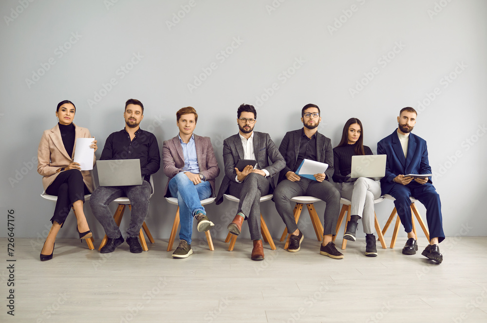 Obraz premium Young professional business people sitting on the chairs in row and looking at the camera. Group of a happy staff. Job candidates seekers with resumes in hand waiting for interview invitation turn.