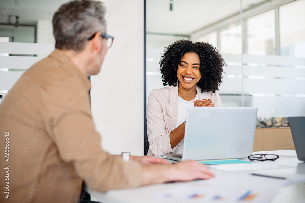 A joyful female professional with curly hair shares a warm smile while engaging in a productive conversation with her mature male colleague in office, depicting an atmosphere of amicable collaboration A joyful female professional with curly hair shares a warm smile while engaging in a productive conversation with her mature male colleague in office, depicting an atmosphere of amicable collaboration