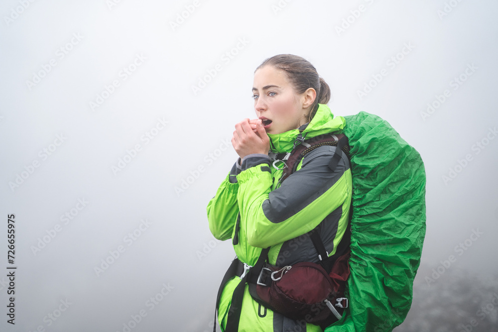 Young mountaineer woman breathes on her hands to keep warm at cold autumn day in a foggy weather, traveler warms her hands with a warm breath, frozen hiker heats from cold, low temperature Young mountaineer woman breathes on her hands to keep warm at cold autumn day in a foggy weather, traveler warms her hands with a warm breath, frozen hiker heats from cold, low temperature