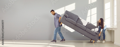 Family couple moving furniture at home. Young man and woman bought sofa and are placing it in living room together. Two happy people moving gray couch inside new house or apartment. Banner background