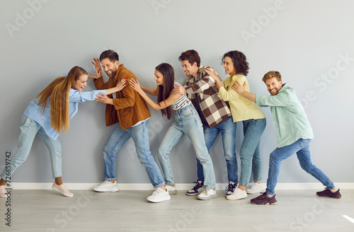 Full length portrait of a funny happy overjoyed group of friends having fun on a gray wall background pushing each other enjoying time together. Team building and friendship concept.