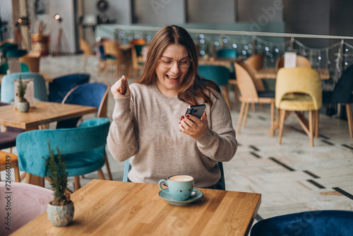 Obraz na płótnie Satisfied young woman with coffee cup holding phone and celebrating in a cafe. hipster female in glasses overjoyed with winning online contest on web site w salonie