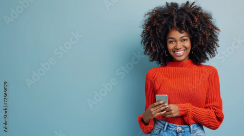 young woman with voluminous curly hair is smiling and holding a smartphone, wearing a red sweater, against a teal background
