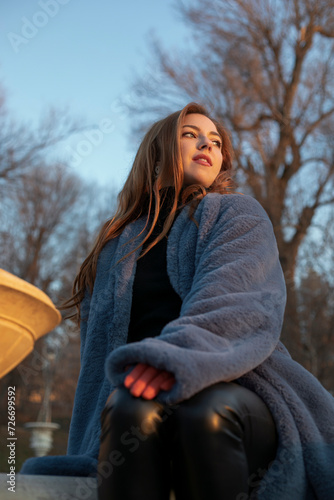 Female beauty portrait, model posing gorgeous in park, dressed in a orange gloves and blue fur coat, with long red curly hair, sunset background.