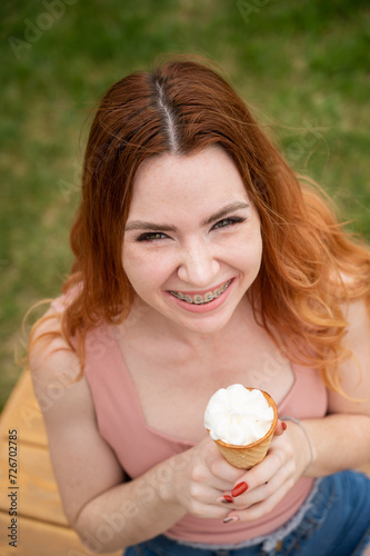 Young beautiful red-haired woman smiling with braces and eating an ice cream cone outdoors. Vertical photo.