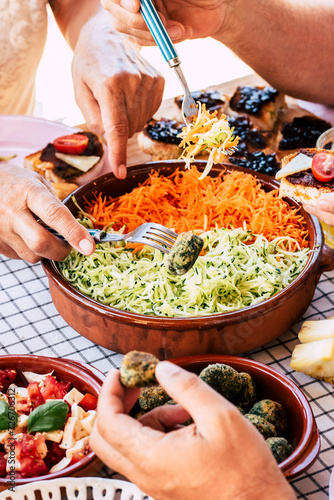 Close up of group of caucasian people take vegetables vegetian food from a table and eat having fun all together at restaurant or home - healthy and colorful food lifestyle