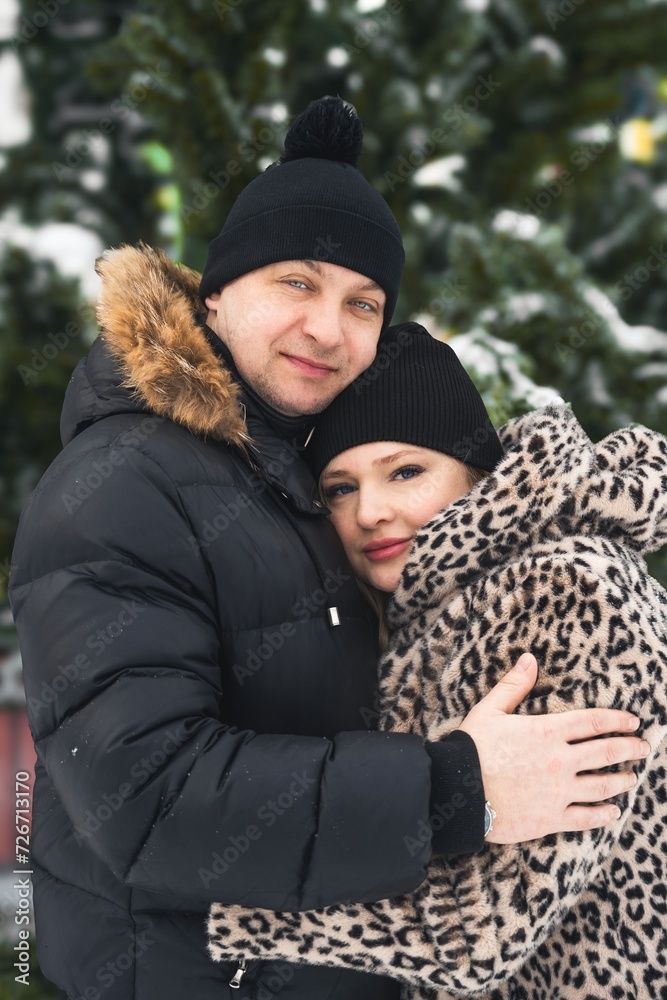 Fototapeta premium Love romantic couple lovestory. Man hugging woman, kissing girl in Christmas winter fur trees decoration. Romantic date. Walking,having fun. Stylish clothes,fur leopard print coat, black jacket, hat
