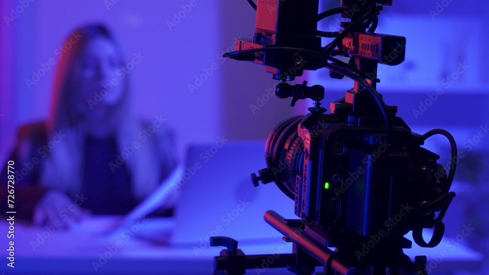 Professional video camera on a tripod against a blurred background of a female presenter in a dark studio illuminated by pink and blue neon lights.