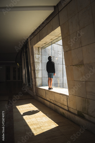 Young man in black hoodie and blue shorts stands in shadowed corridor at Cidade da Cultura in Santiago de Compostela. Sunlight streams through square opening, casting captivating shadow play.