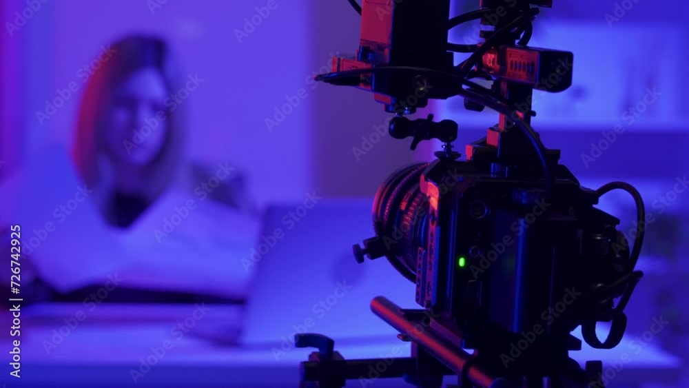 Professional video camera on a tripod against a blurred background of a female presenter in a dark studio illuminated by pink and blue neon lights. The operator's hands adjust the camera, close up. Professional video camera on a tripod against a blurred background of a female presenter in a dark studio illuminated by pink and blue neon lights. The operator's hands adjust the camera, close up.
