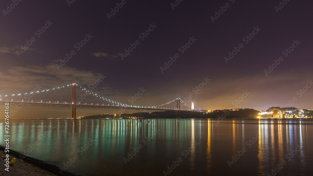 Fototapeta premium Illuminated red bridge 25 de Abril Bridge crossing the Tagus river with statue of Cristo Rei during night time, Lisbon, Portugal